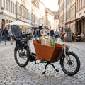 Hebie Zweibeinständer lite REX am Lastenrad mit Kindersitz und Einkauf in belebter Gasse.