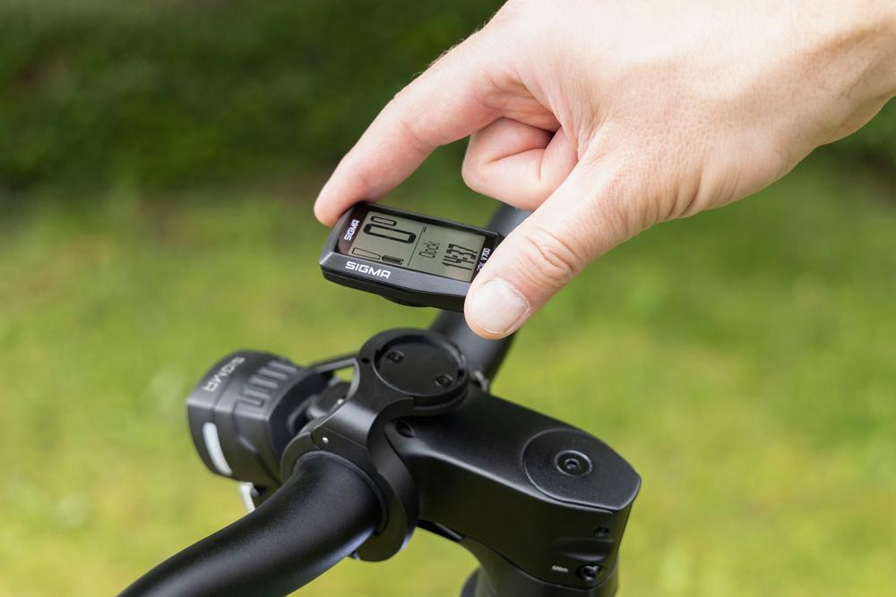 Hand hält schwarzen Sigma EOX VIEW 700 E-Bike-Computer über Fahrradlenker-Halterung.