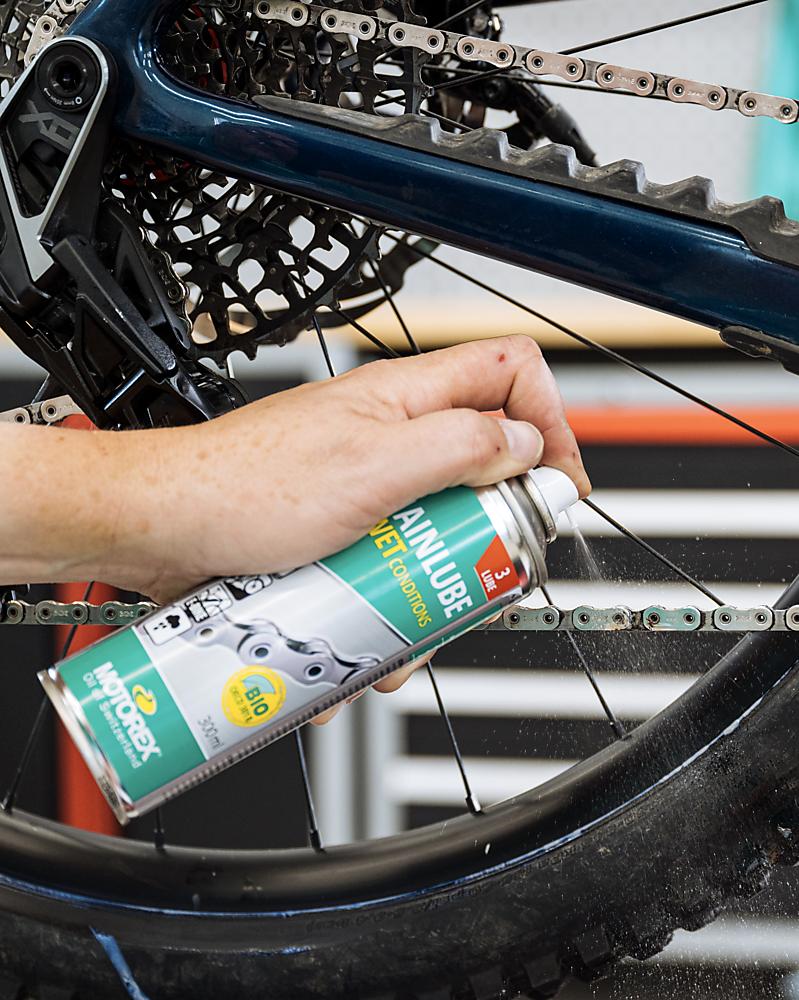 Hand sprüht MOTOREX CHAINLUBE WET CONDITIONS Nassschmierstoff auf Fahrradkette für Regen und Matsch.