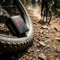 Specialized SV MTB-Fahrradschlauch (Box) am Reifen auf staubigem Trail, Mountainbiker im Hintergrund.