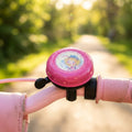 Rosa Lillifee Fahrradklingel mit Motiv auf pinkem Kinderfahrrad, Wassertropfen, sonniger Weg im Hintergrund.