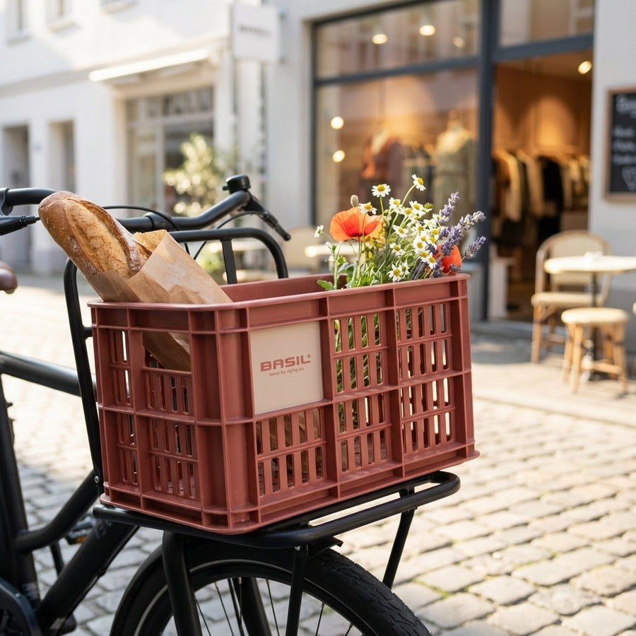 Basil Crate S Fahrradkiste 17,5L terra red auf Gepäckträger mit Baguette und Blumen in einer Stadtstraße.
