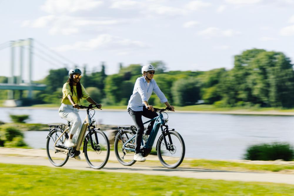 Zwei Personen radeln auf City-E-Bikes am Flussufer. Schwalbe Motion Big Apple 29 Zoll Komfort-Drahtreifen.