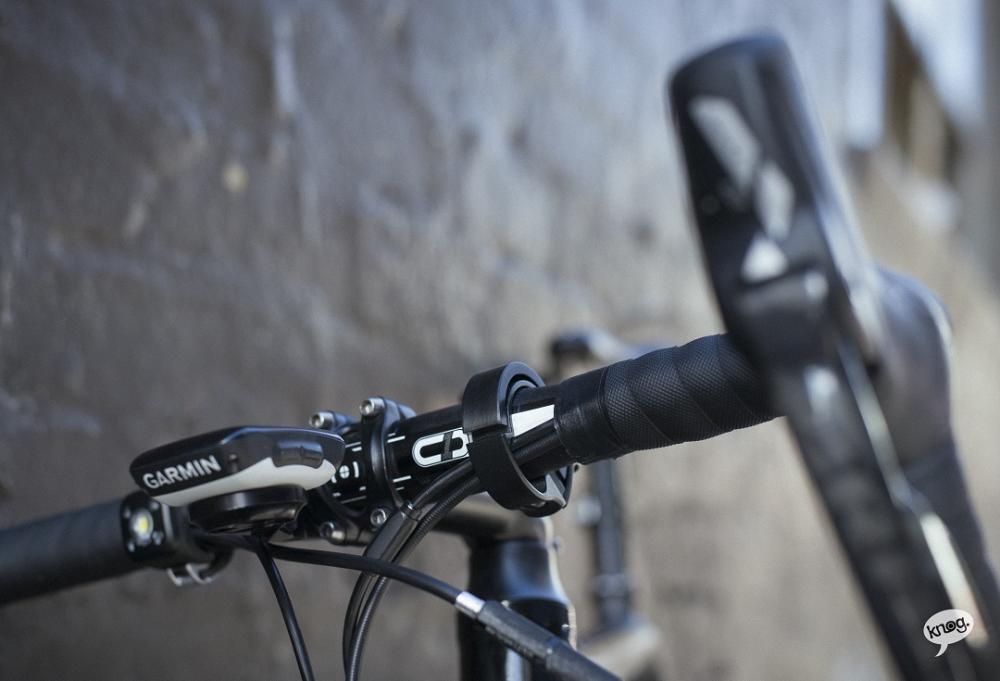 Knog Oi Classic Large Fahrradklingel in Schwarz am Lenker neben einem Garmin-Fahrradcomputer montiert.